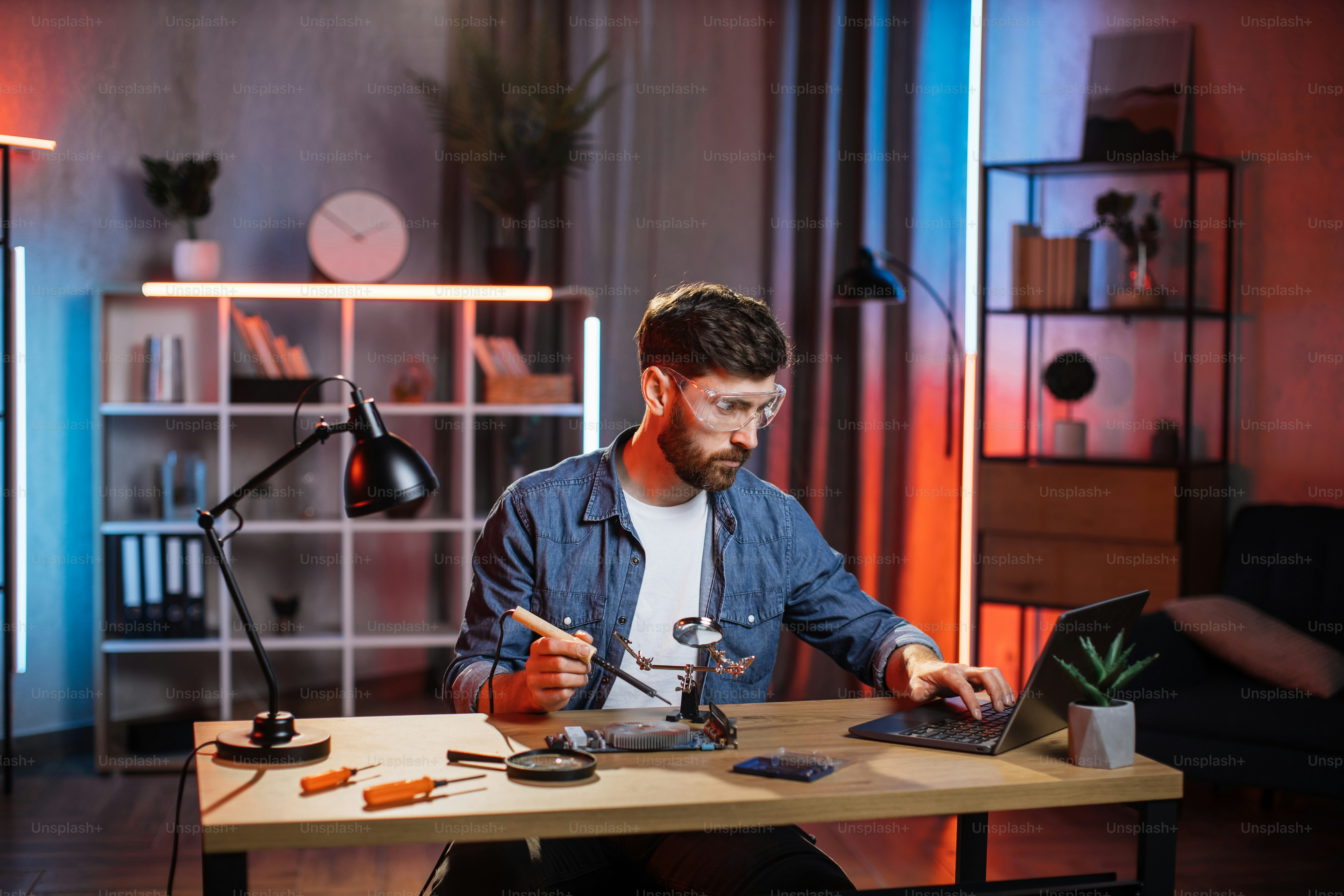 Young man in casual wear surfing internet on laptop for searching way of repairing broken video card. Bearded guy sitting at table, holding soldering iron and looking on computer screen.