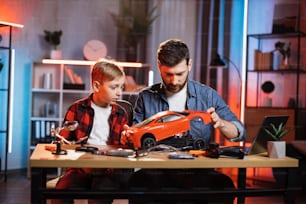 Focused bearded man and little boy sitting together at table and examining broken remote controlled car. Young caucasian father helping his son to fix favorite toy at home.