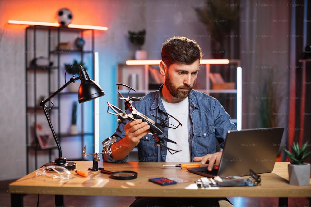 Handsome bearded man using wireless laptop for searching for solution of fixing modern quadcopter. Concept of people, technology and repairing.