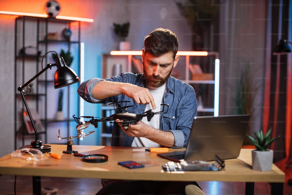 Concentrated bearded man using various tools for repairing modern quadcopter at home. Young caucasian guy in denim shirt fixing flying drone by himself.