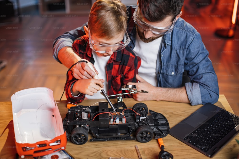 Father with son in protective glasses using soldering iron for repairing red toy car at home. Concept of maintenance, family and togetherness.