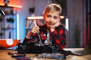 Portrait of caucasian boy in casual outfit soldering broken remote controlled car at home. Cute child sitting at table and repairing favorite toy.