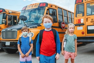 Three children kids students in protective face masks near school yellow bus outdoors. New normal at coronavirus covid-19 pandemic. Measurements against virus spread in class.