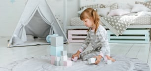 Beautiful little girl wearing pajamas playing with wooden toy blocks in the kids room. Natural eco toys concept. Selective focus.