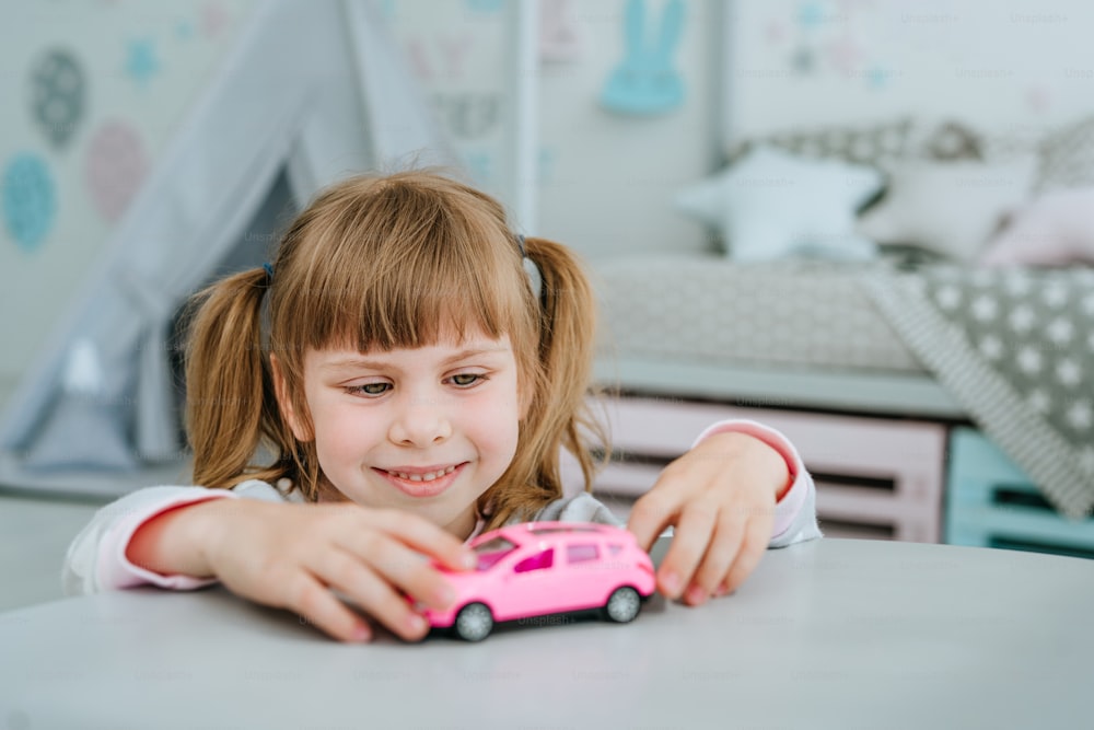 Hermosa niña en pijama jugando con un coche de juguete rosa en la habitación de los niños. Enfoque selectivo.