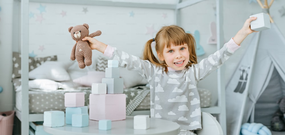 Beautiful little girl wearing pajamas playing with wooden toy blocks and bear toy sitting at the table in the kids room. Natural eco toys concept. Selective focus.