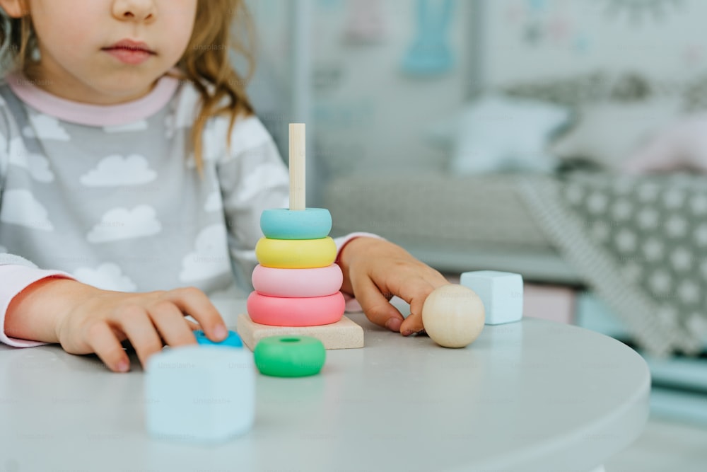 Close up of little toddler girl wearing pajamas playing with wooden toy pyramid sitting at the table in the kids room. Natural eco toys concept. Selective focus.