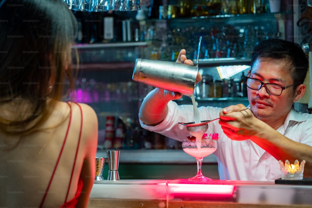 Selbstbewusste asiatische Frau, die an der Bartheke sitzt, alkoholische Getränke trinkt und sich mit einem Barkeeper im Nachtclub unterhält. Männlicher Barkeeper bereitet Alkoholgetränk für Kunden zu. Small Business Bar und City Nightlife Konzept