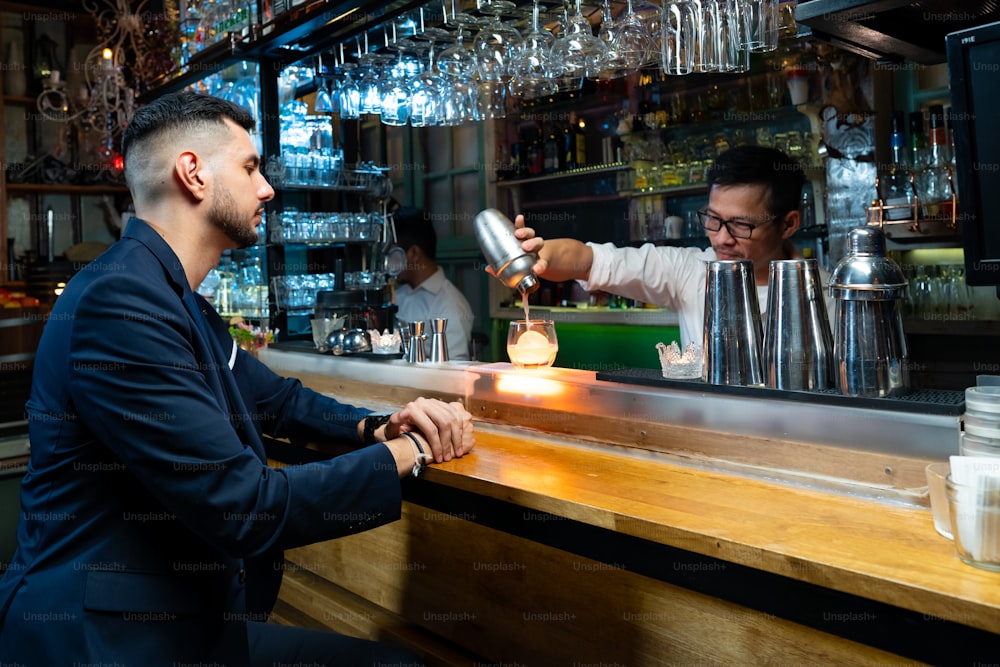 Vertrauen kaukasischer Mann, der an der Bartheke sitzt, genießt es, auf alkoholische Getränke zu warten und mit Barkeeper im Nachtclub zu sprechen. Männlicher Barkeeper bereitet Alkoholgetränk im Shaker mit Eisball auf Steinglas zu.