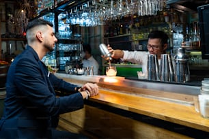 L'uomo caucasico seduto al bancone del bar si diverte ad aspettare una bevanda alcolica e a parlare con il barista in discoteca. Barista mixologist maschio che prepara una bevanda alcolica in uno shaker con palla di ghiaccio su vetro rock.
