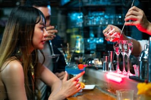 常夜灯のあるナイトクラブのバーカウンターでカクテルグラスに酒とシロップを注ぐプロのアジア人男性バーテンダー。ミクソロジストのバーテンダーが、ミックスアルコールのファンシーカクテルドリンクをお客様に提供しています。