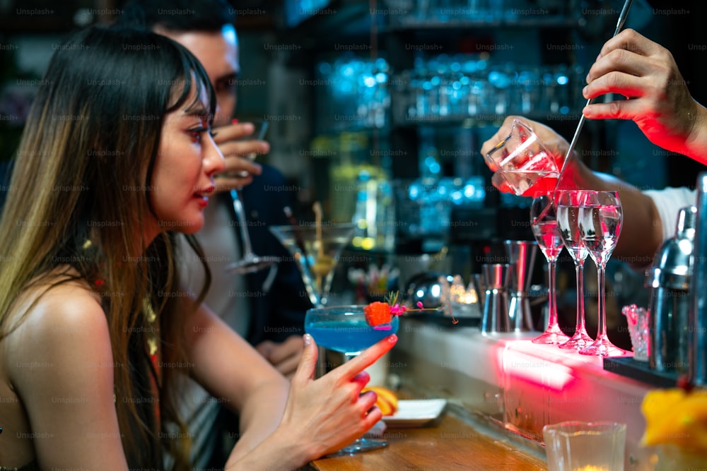 Professional Asian male bartender pouring liquor and syrup into cocktail glass on bar counter at nightclub with night lights. Mixologist barman making mixed alcoholic fancy cocktail drink serve to customer.