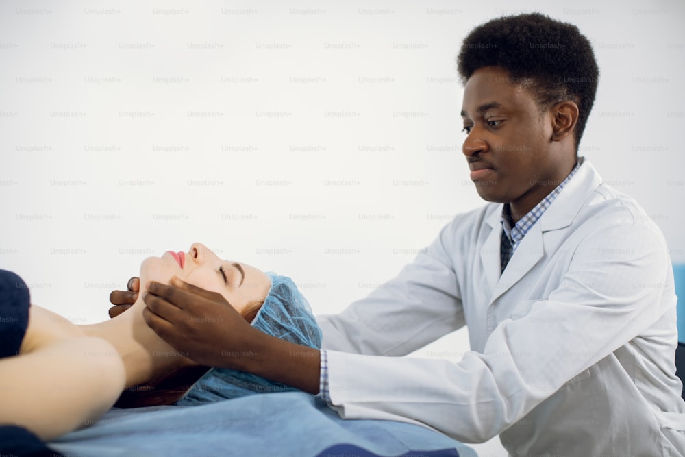 Handsome young professional African American man masseur of medical center, conducting anti-aging treatment facial massage for young pretty female client, lying on massage table