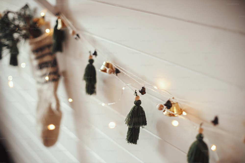 Stylish christmas garland with bells, rustic stocking with fir branch and christmas lights on wooden wall. Festive decorated atmospheric scandinavian room. Boho handmade decor. Space for text