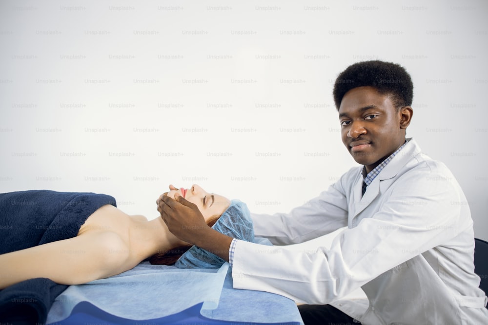 Handsome young professional African American man masseur of medical center, conducting anti-aging treatment facial massage for young pretty female client, lying on massage table