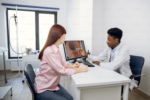 Confident African American male doctor in his modern office, explaining results of endoscopy to young female Caucasian patient, pointing on the image of digestive tract on computer