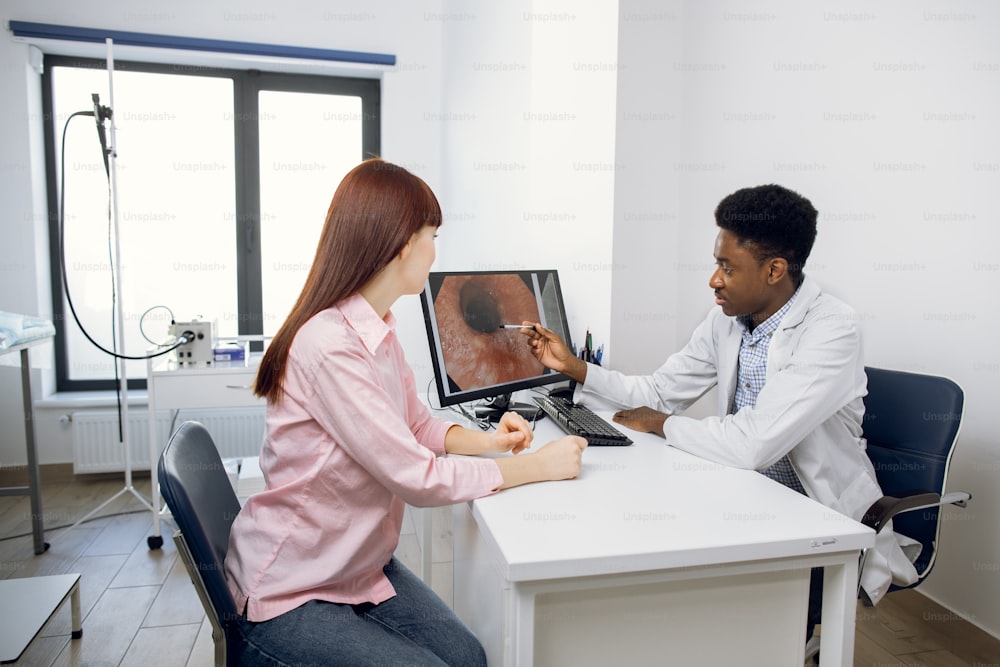 Confiante médico afro-americano do sexo masculino em seu escritório moderno, explicando os resultados da endoscopia para jovem paciente caucasiano do sexo feminino, apontando para a imagem do trato digestivo no computador