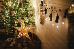 Stylish Christmas star and tree decorated with modern white baubles, boho ornaments and golden lights in atmospheric evening room. Festive scandinavian room at eve. Magic time. Space for text