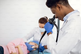 Retrato de primer plano de un médico afroamericano de 30 años mirando en la cámara del endoscopio, haciendo endoscopia de la paciente femenina junto con la enfermera.