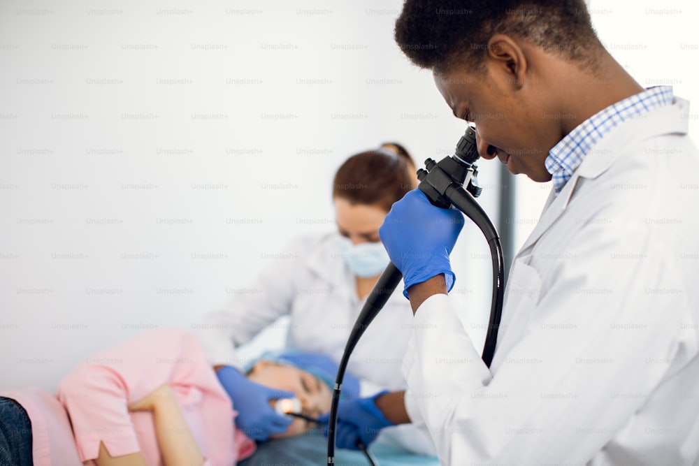 Close up portrait of 30-aged Afro-American man doctor looking in endoscope camera, doing endoscopy of female patient together with nurse.