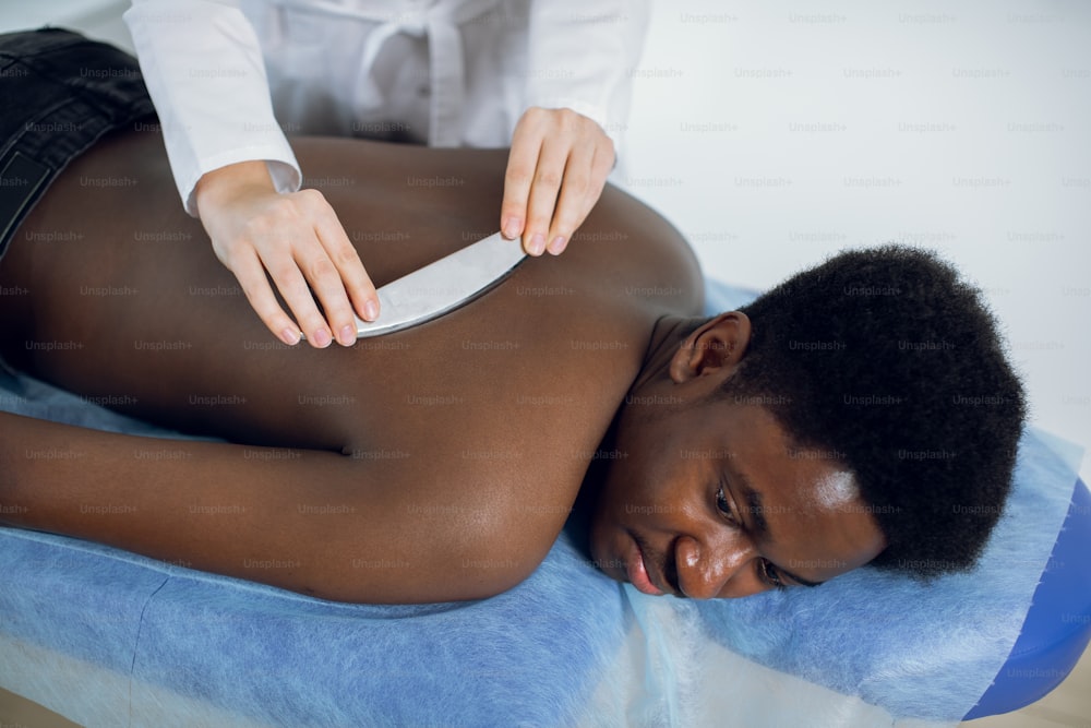 Top angle view of relaxed likable African American man lying on the couch while female massage therapist is massaging his back using steel metal Gua Sha scraper device