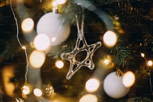 Modern glass christmas ornaments, wooden garland, white baubles and golden christmas lights close up. Atmospheric festive decorated scandinavian room. Stylish christmas decor, glass star