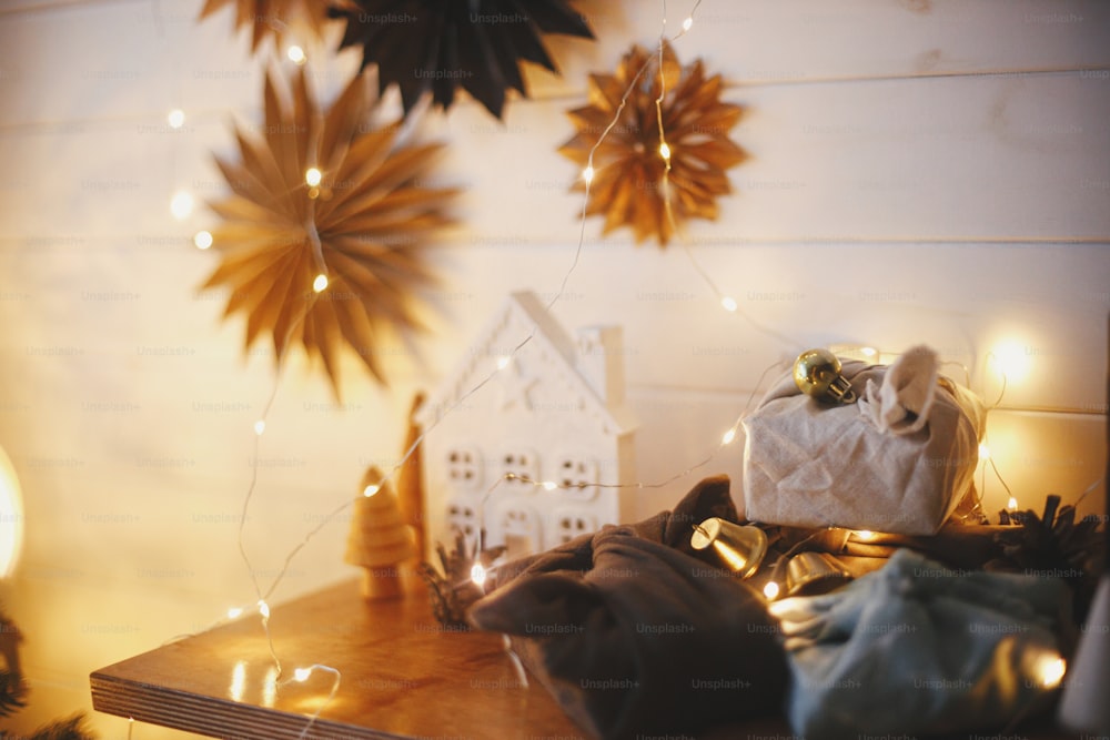 Merry christmas! Stylish plastic free christmas gifts on wooden shelf on background of little house, trees, paper stars and christmas golden lights bokeh. Atmospheric festive scandinavian room