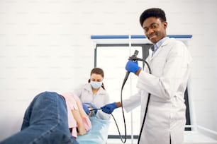 African male doctor endoscopist surgeon in medical suit, and gloves holds an endoscope, posing on camera, ready for endoscopy procedure for female patient