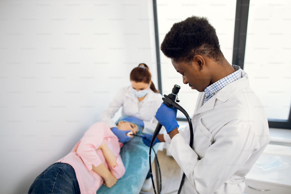 内視鏡を持つアフリカ系アメリカ人の男性医師と、若い女性患者を診察する女性の同僚看護師の接写。内視鏡検査、健康診断