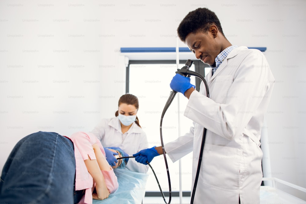 Young african american man doctor and female nurse doing endoscopy procedure for female patient, lying on the couch in modern examination room in clinic