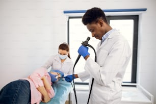 Examen endoscópico en la clínica médica moderna. Joven médico afroamericano seguro de sí mismo sostiene un endoscopio en la mano, insertando la cámara en la boca de la paciente