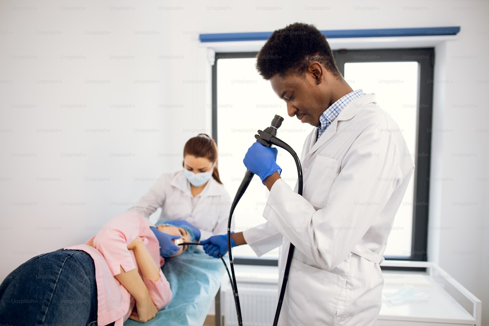 Esame endoscopico in clinica medica moderna. Il giovane medico maschio afroamericano fiducioso tiene un endoscopio in mano, inserendo la macchina fotografica nella bocca del paziente femminile