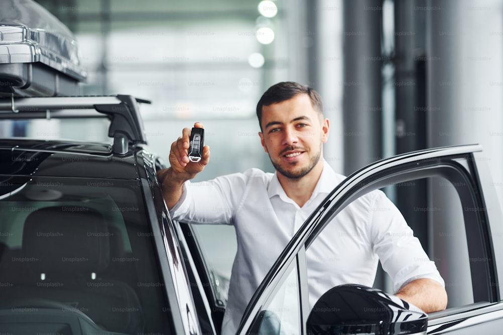 Holds keys. Young man in white shirt is indoors with modern new automobile.