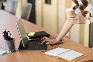 Primer plano, vista lateral, mujer de negocios se apoya en el escritorio de la oficina, escribiendo en el teclado de la tableta mientras sostiene el café para llevar en la oficina