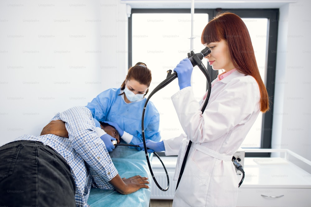 女性集中医師胃腸病専門医、ソファに横たわっている若いアフリカ系アメリカ人男性患者の胃内視鏡手術中に内視鏡を操作します