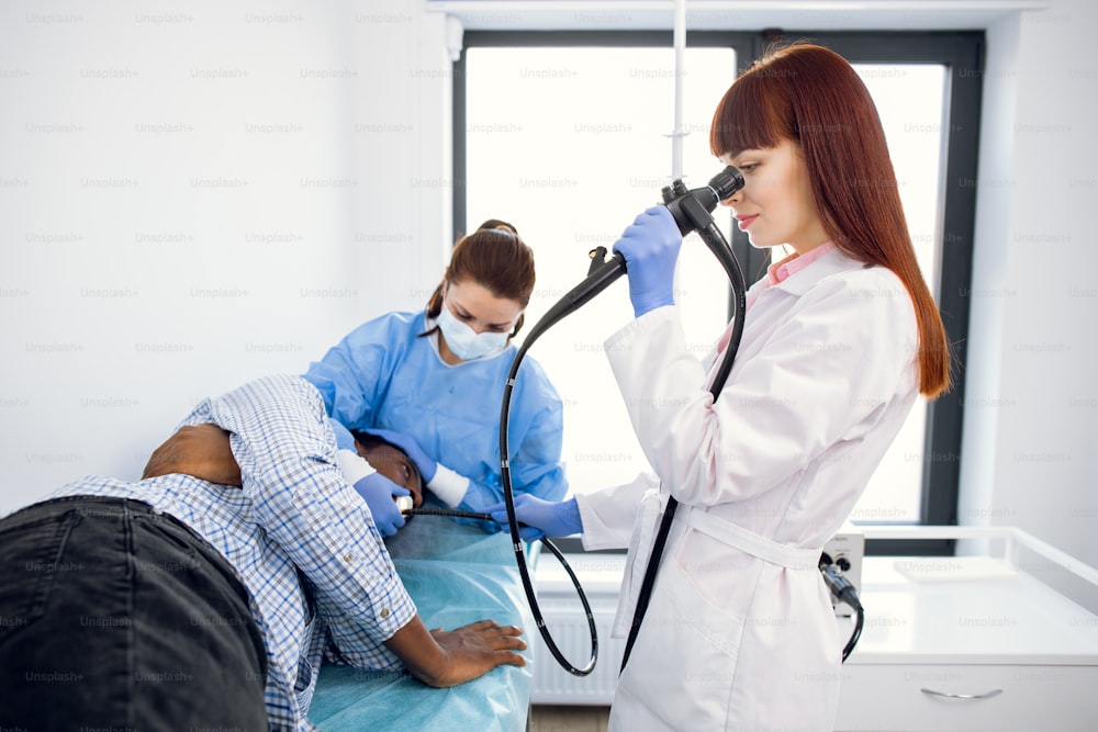 Female concentrated doctor gastroenterologist, operating endoscope during gastroscopic procedure of young afro-american man patient, lying on the couch