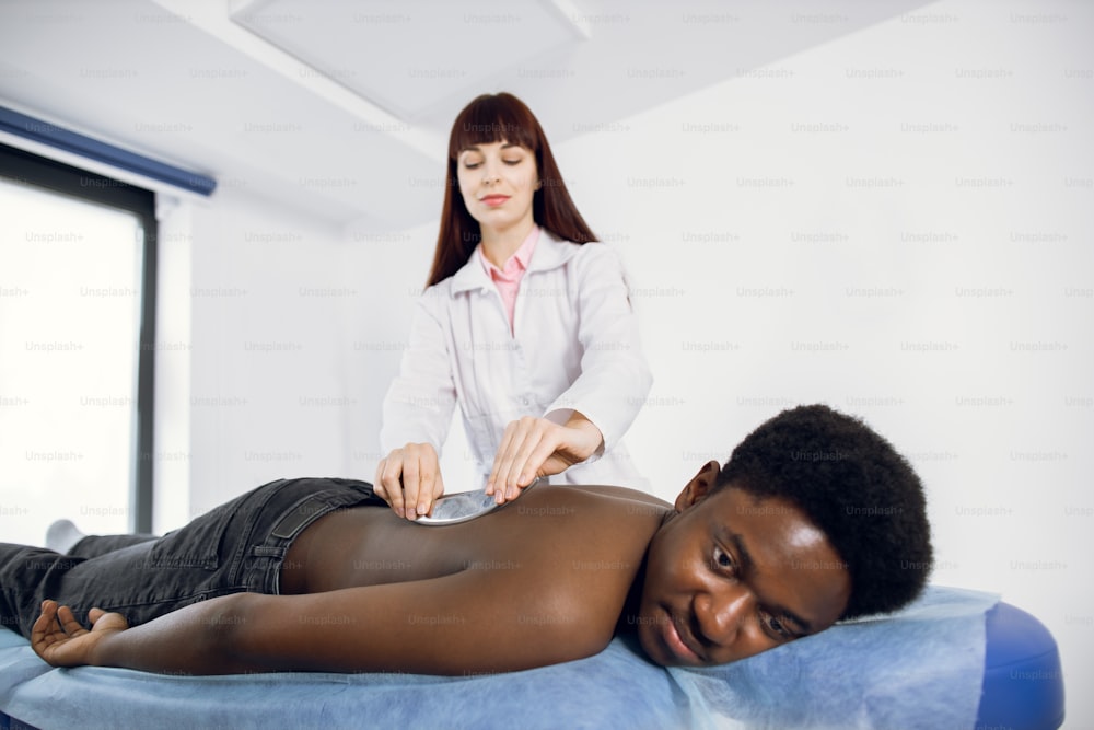 Back massage using medical steel metal massager scraper. Young confident woman doctor doing Gua Sha massage for handsome African man patient with back pain