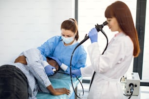 Gastroenteróloga médica concentrada femenina, endoscopio operativo durante el procedimiento gastroscópico de un joven paciente afroamericano, acostado en el sofá