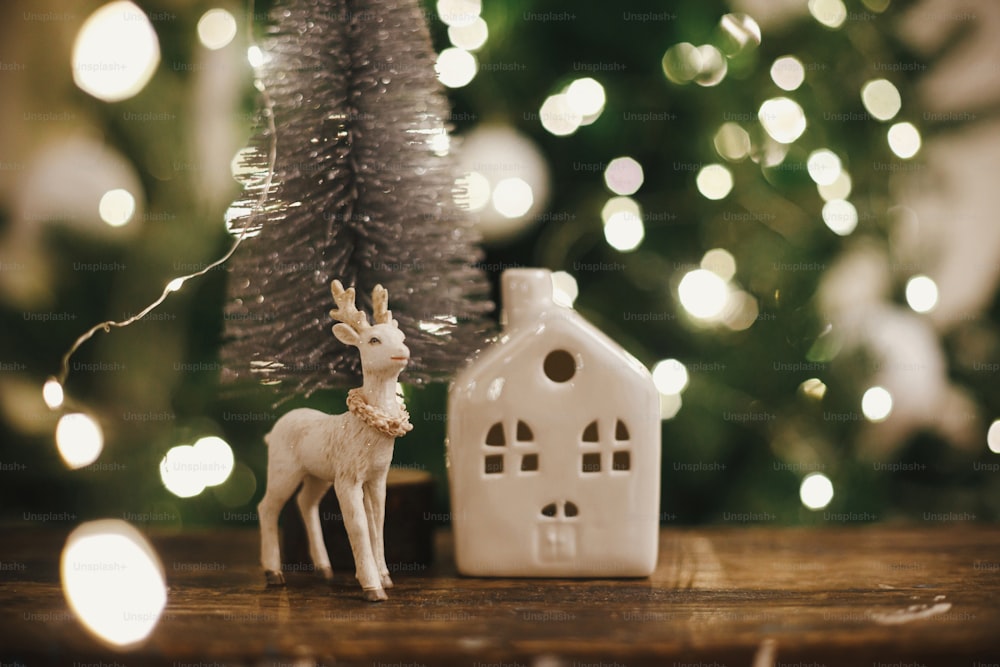 Stylish christmas tree, reindeer toy and house on rustic wood on background of christmas lights. Beautiful little christmas scene. Merry christmas. Magical christmas time. Space for text