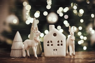 Stilvolles Rentierspielzeug, Weihnachtsbäume und Haus auf rustikalem Holz vor dem Hintergrund der Weihnachtsbeleuchtung. Schöne moderne kleine Weihnachtsszene. Frohe Weihnachten und frohe Festtage! Magisches Winterdorf