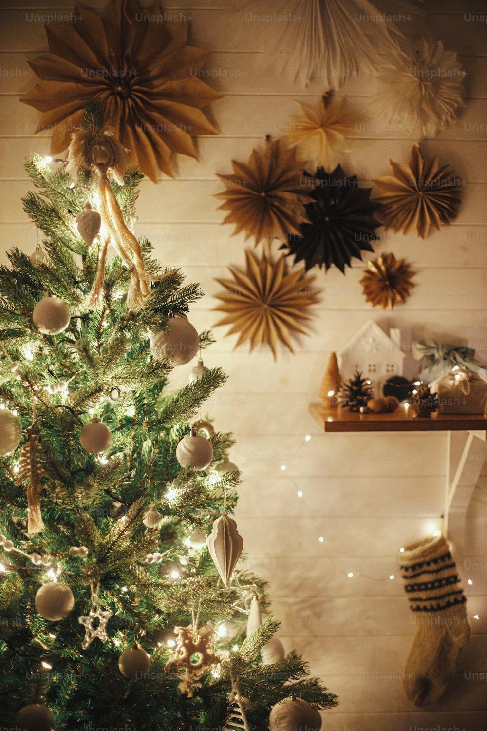 Stylish Christmas tree decorated with modern white baubles, boho ornaments and golden lights on background of paper stars on wall and stocking. Atmospheric festive scandinavian room. Magic time