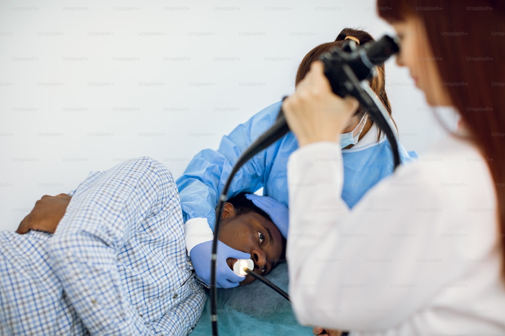 Jeune patient afro-américain allongé sur le canapé à l’hôpital, pendant l’endoscopie, tandis que l’équipe de deux femmes médecins caucasiennes effectue la procédure