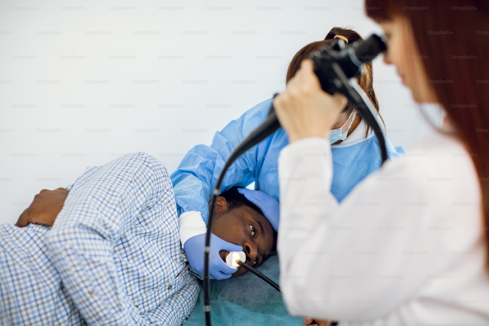 Young African American man patient lying on the couch in hospital, during endoscopy, while team of two Caucasian female doctors conducting procedure