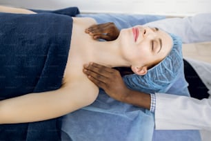 Close up top view of young relaxed lady, lying on the couch and enjoying massage. Hands of male Afro-American doctor masseur cosmetologist makes a decollete massage procedure.