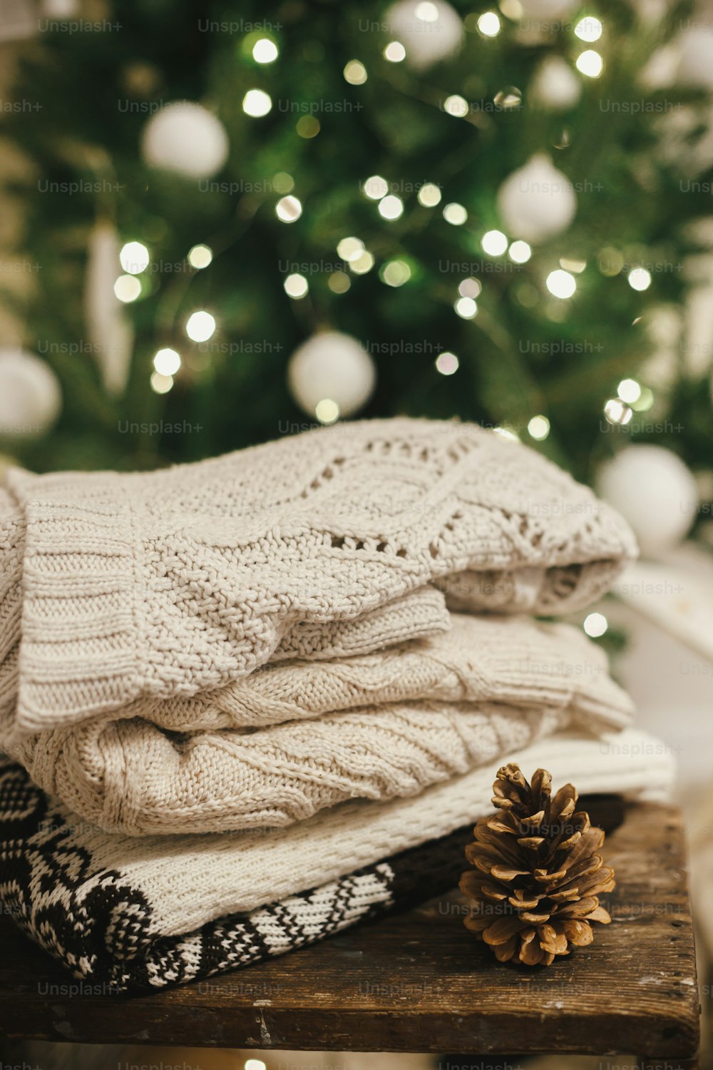 Cozy knitted sweaters on rustic wood with pine cone on background of christmas tree in lights and white star. Pile of stylish sweaters in festive decorated scandinavian room. Winter holidays