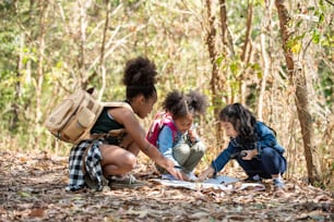 夏の晴れた日に森の山で一緒にハイキングするバックパックを持つ多様性の小さな女の子の友達のグループ。座って地図を眺めながら森を探検する3人の子供たちが野外活動を楽しんでいます。