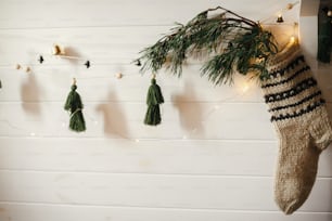 Weihnachtslicher rustikaler Strumpf mit Tannenzweig und stilvoller Girlande mit Glocken und Bäumen an weißer Holzwand mit Weihnachtsbeleuchtung. Festlich dekoriertes stimmungsvolles skandinavisches Zimmer. Modernes handgefertigtes Dekor
