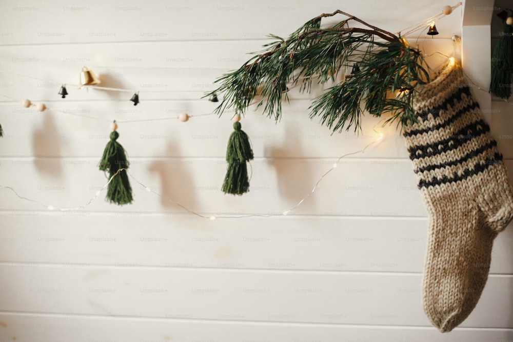 Christmas rustic stocking with fir branch and stylish garland with bells and trees on white wooden wall with christmas lights. Festive decorated atmospheric scandinavian room. Modern handmade decor