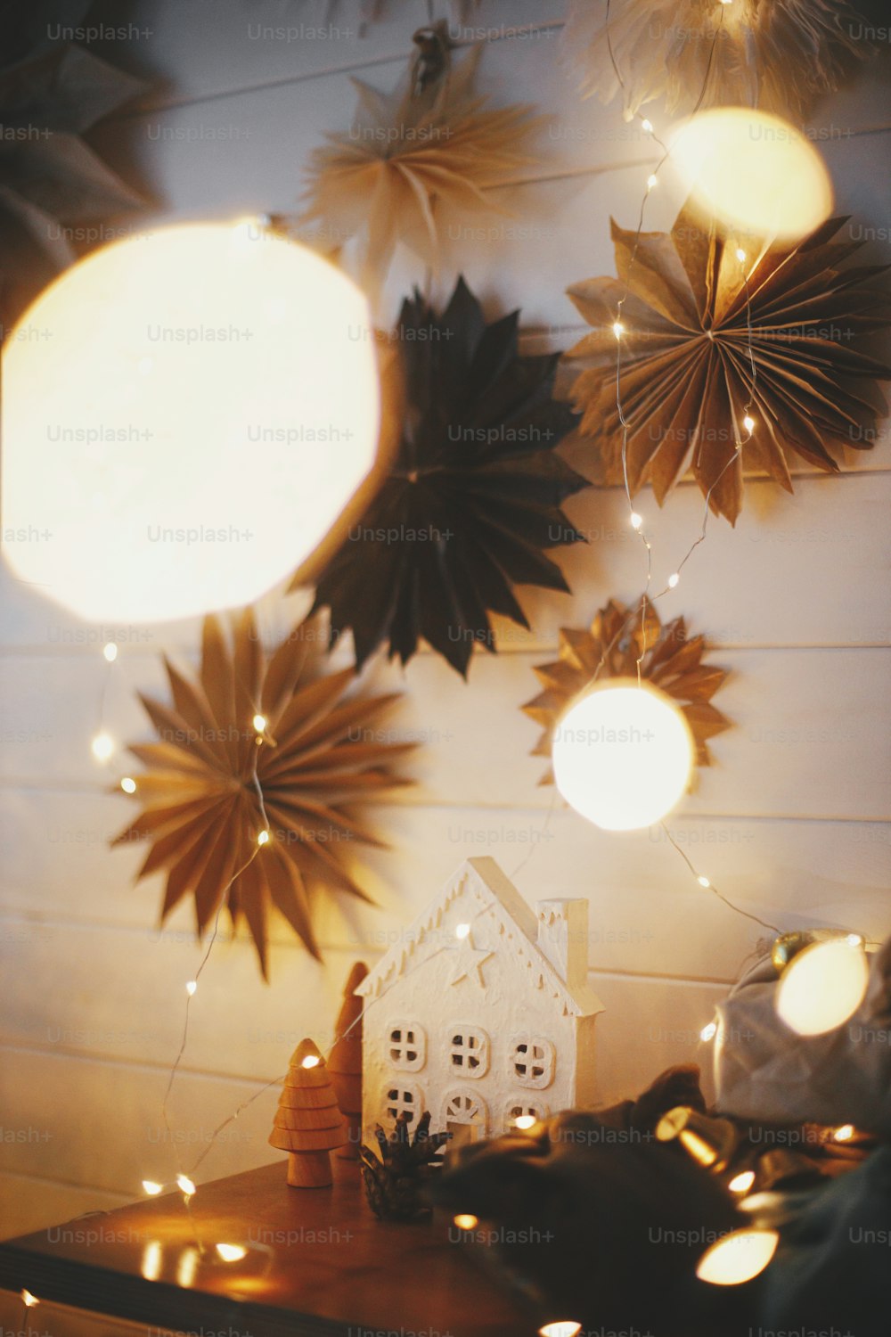 Stylish christmas little house and trees, plastic free gifts on wooden shelf on background of white wall with paper stars and golden lights. Festive decorated scandinavian room. Atmospheric eve