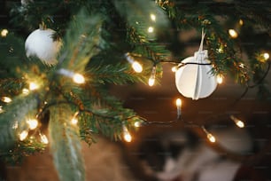 Modern boho christmas tree ornaments, white baubles, wooden garland, christmas lights close up. Atmospheric festive decorated scandinavian room. Stylish christmas decor